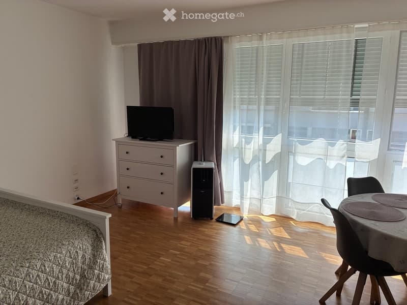A well-lit room with wooden flooring, a bed, a dresser with a TV, a small table with chairs, and large windows with curtains.