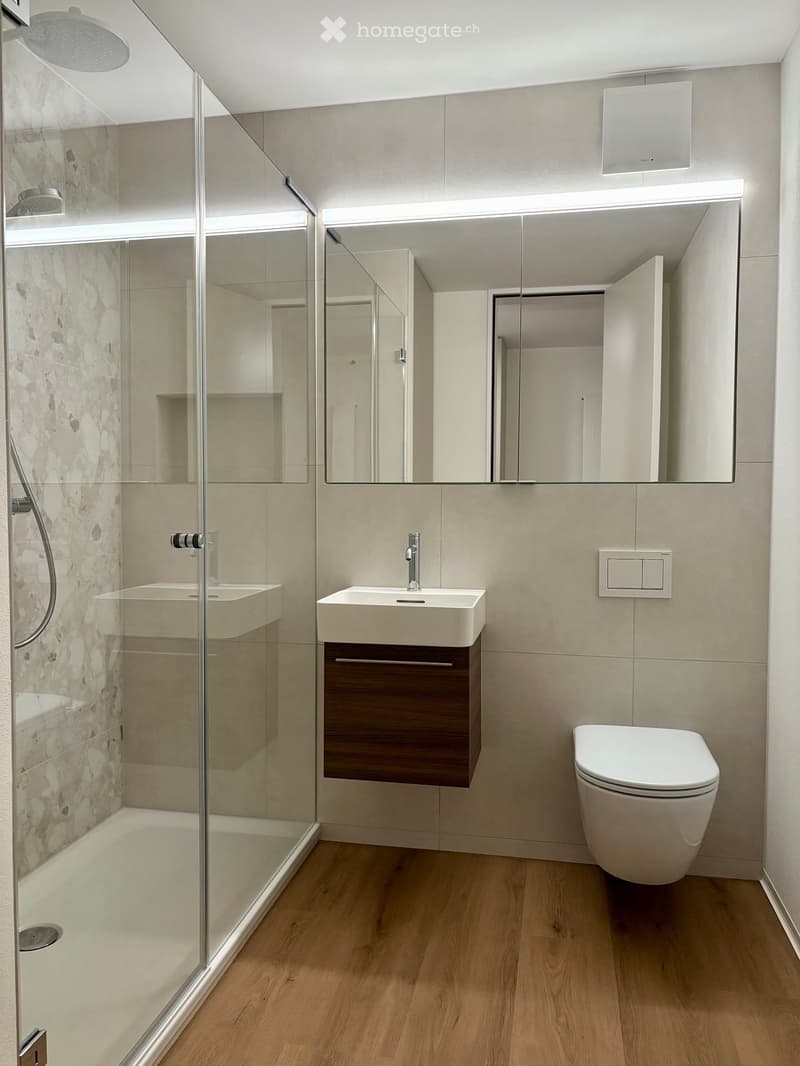 Modern bathroom with glass shower enclosure, marble tiles, white sink, wooden vanity, large mirror, recessed lighting