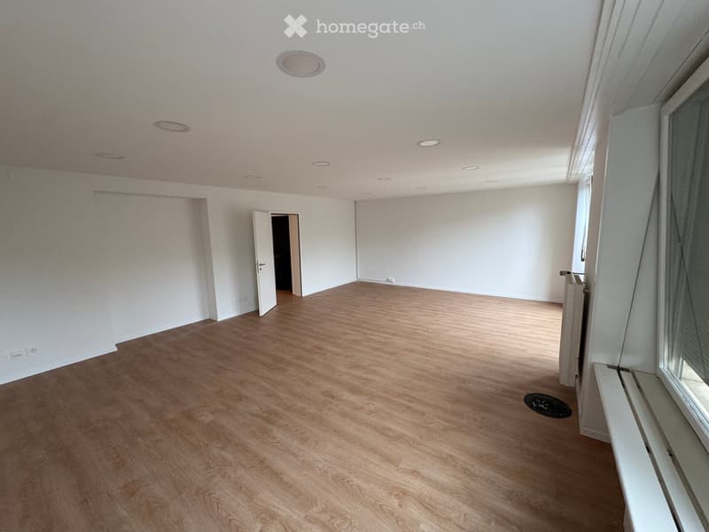 Empty room with wooden flooring, white walls, and recessed ceiling lights. Features a large window with a sill and an open doorway leading to another space.