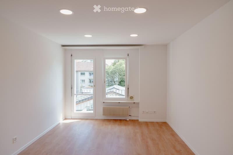 Empty room with light wooden flooring, white walls, and large windows offering a view of the outside. The room features recessed ceiling lights and a radiator under one window.
