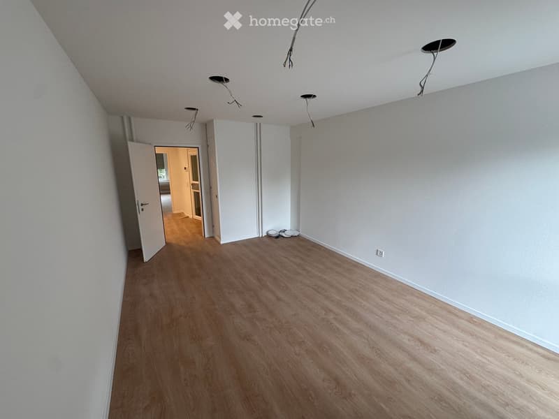 Empty room with light-colored walls, wooden flooring, and several ceiling spotlights. An open doorway leads to another room with similar flooring.