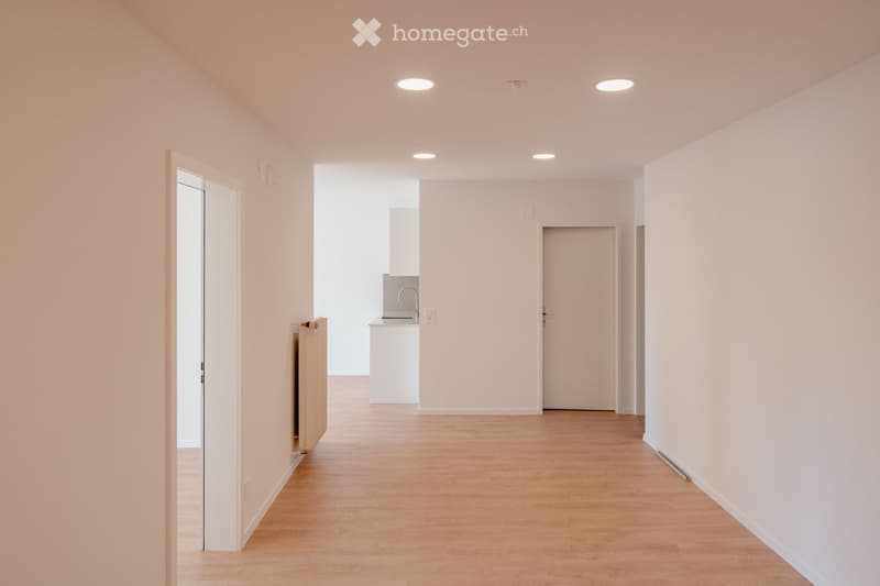 Empty hallway with light wooden flooring, white walls, recessed ceiling lights, and doors leading to adjacent rooms.