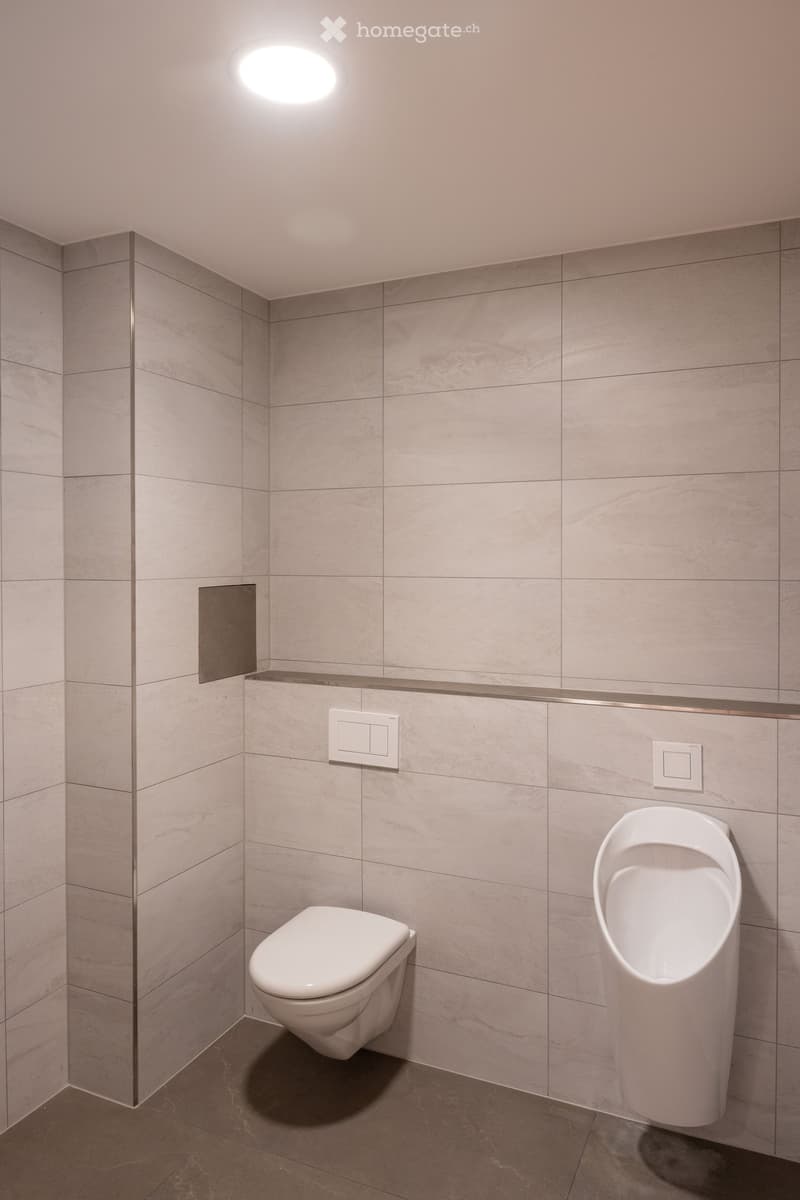 Modern bathroom with a toilet and urinal, tiled walls and floor, and recessed lighting.