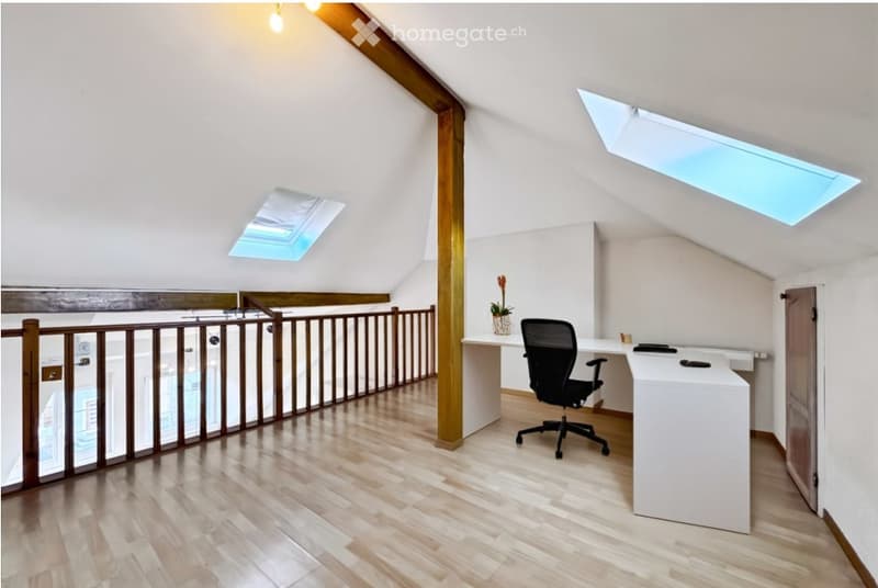 Bright, modern attic room with wooden flooring, white walls, and skylights. Features a white desk, black office chair, and a small flower pot on the desk.