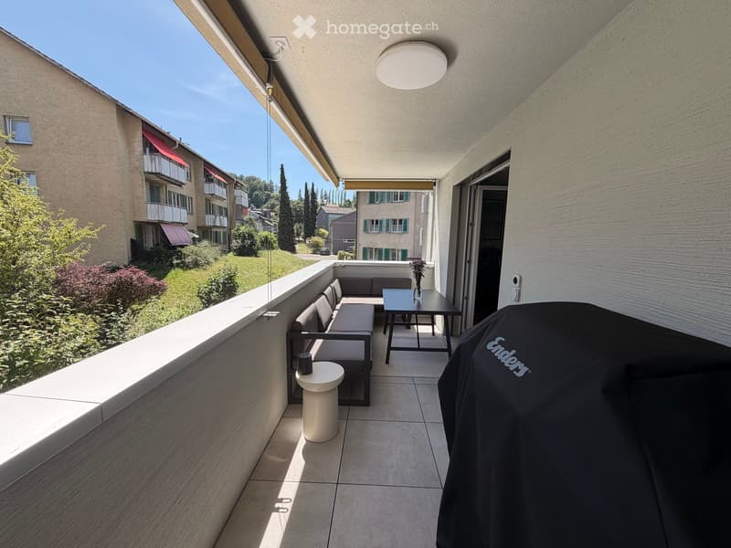 Balcony with a sofa, small table, and stool, overlooking a green area with buildings and trees.