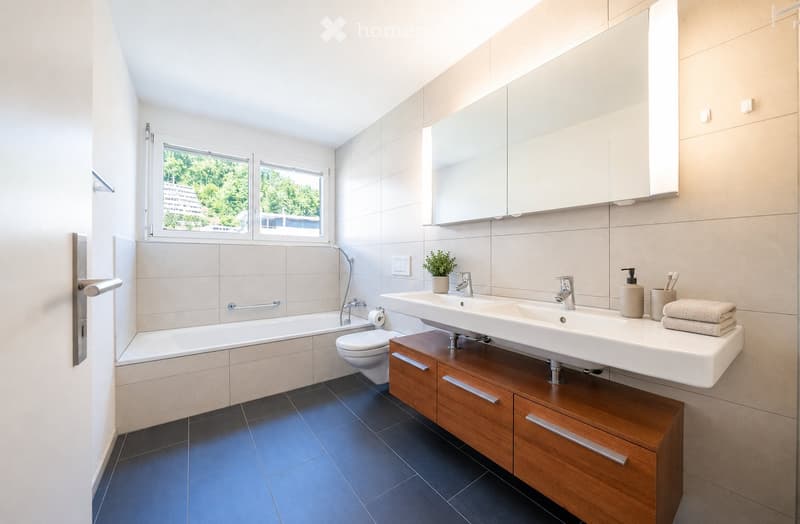 Modern bathroom with a bathtub, toilet, double sink, large mirror, wooden cabinet, and a small plant on the sink counter.
