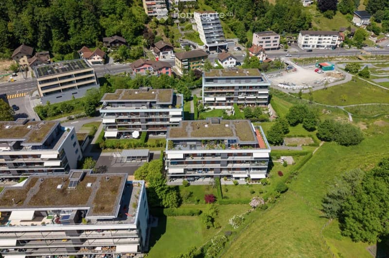 Aerial view of a residential area with multiple apartment buildings, green roofs, and surrounding greenery. The buildings are multi-story with balconies and modern architecture.