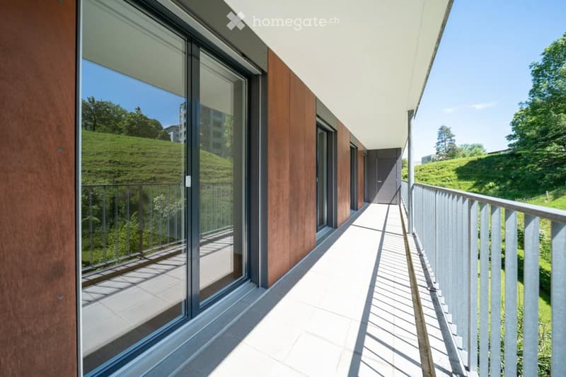 Modern balcony with large glass windows, wooden paneling, metal railing, and a view of a green hillside.
