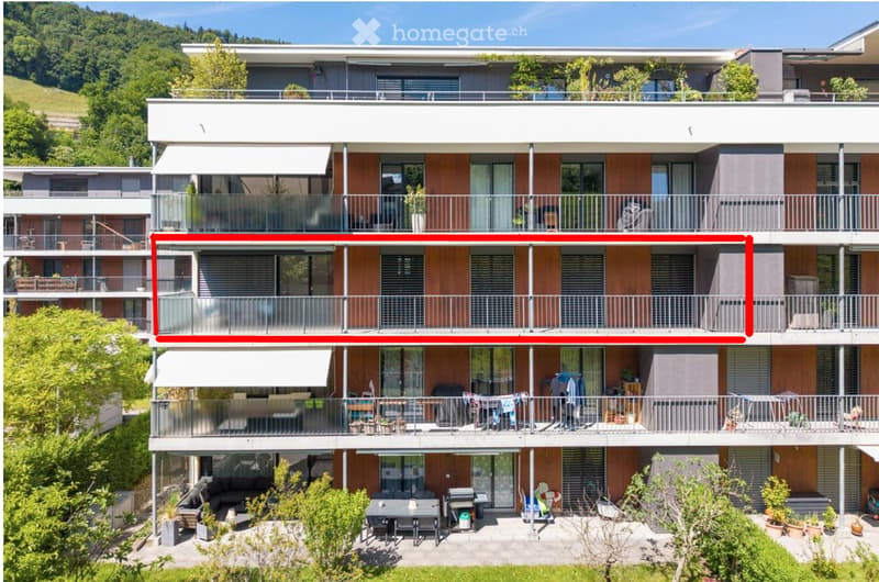 Modern multi-story residential building with balconies, large windows, and contemporary design. Features include wooden paneling, metal railings, and a scenic hillside backdrop.