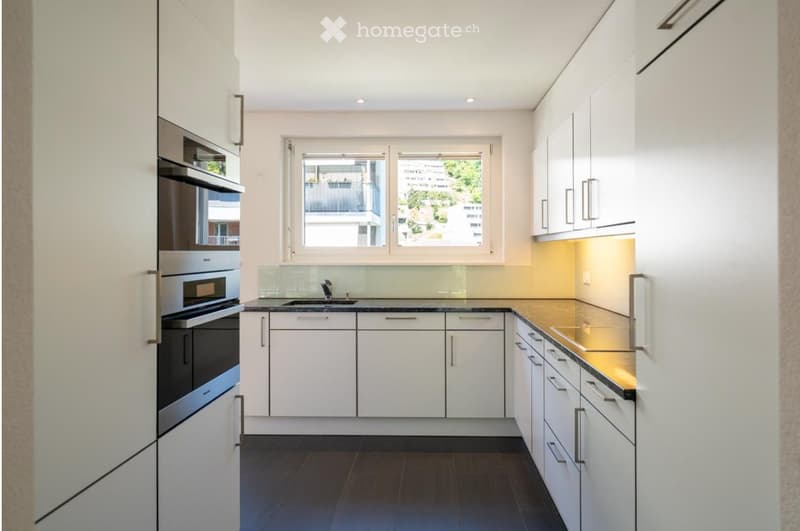 Modern kitchen with white cabinets, stainless steel oven, sink, and a window overlooking a balcony.