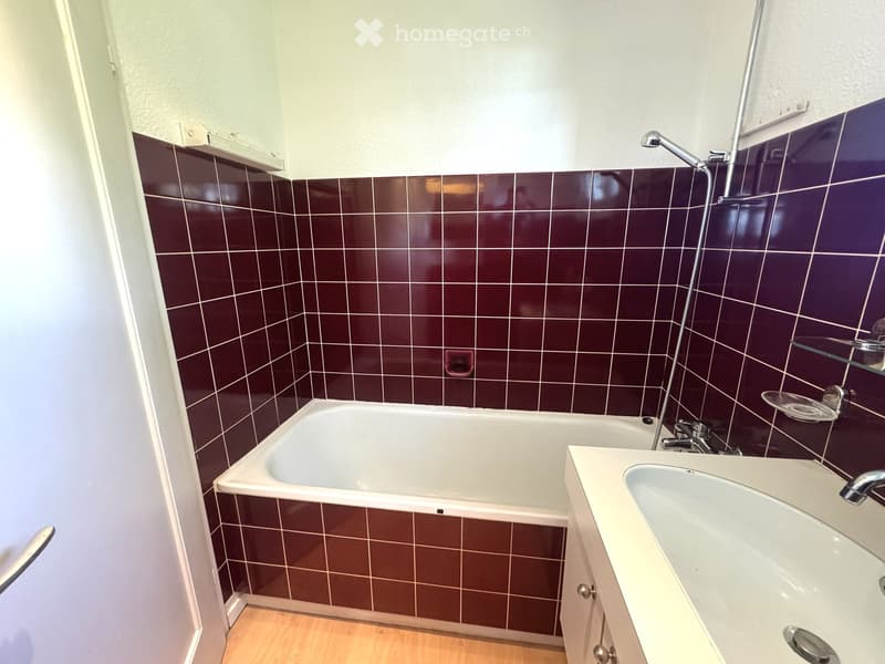 Bathroom with a bathtub, sink, and glass shelf. Dark brown tiles on walls, light-colored wooden floor.