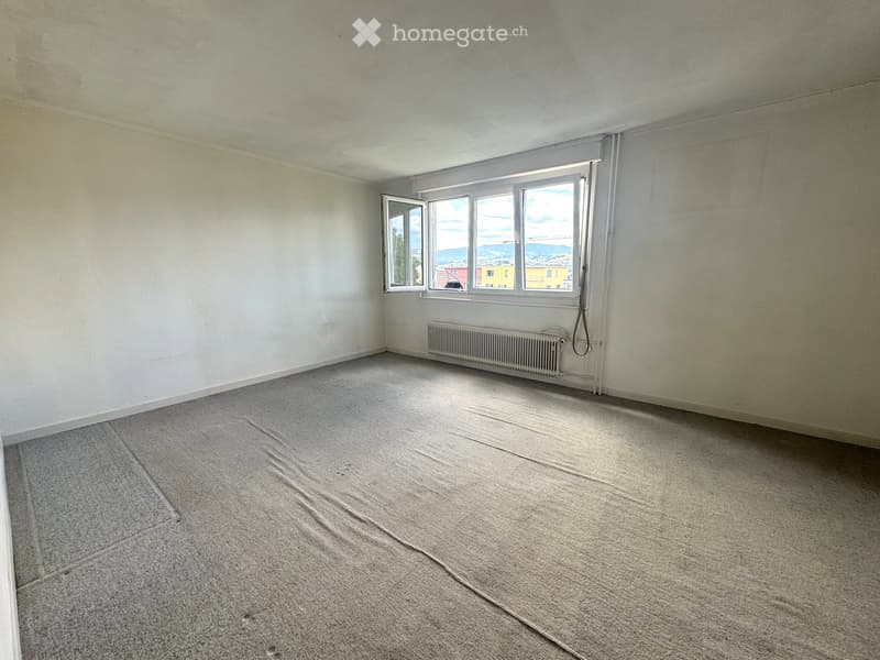 Empty room with light-colored walls, carpeted floor, and a window with a view of the outside. A radiator is visible below the window.