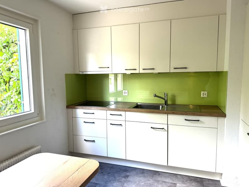 Modern kitchen with white cabinets, wooden countertops, and a green backsplash. Features a sink with a modern faucet, multiple drawers, and a window with a view of greenery.