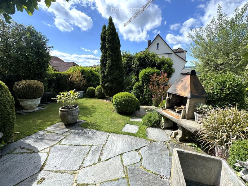 Well-maintained garden with a stone pathway, neatly trimmed hedges, potted plants, a stone bench, and a small outdoor fireplace.