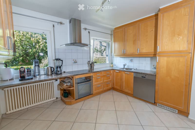 Bright kitchen with wooden cabinets, granite countertop, stainless steel appliances including a coffee maker, blender, oven, and dishwasher. Natural light from windows, tiled backsplash, and tiled floor.