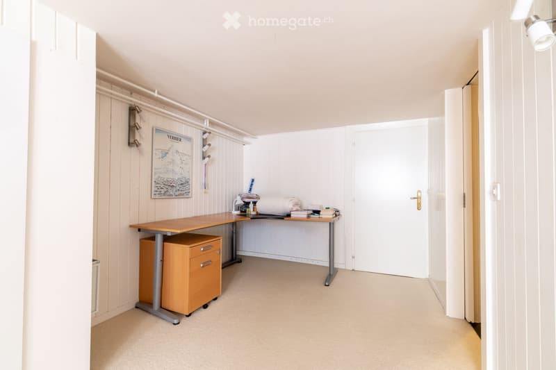 A well-lit, furnished room with a desk, chair, filing cabinet, and a picture frame on the wall. The room features white walls, a carpeted floor, and a door leading to another area.