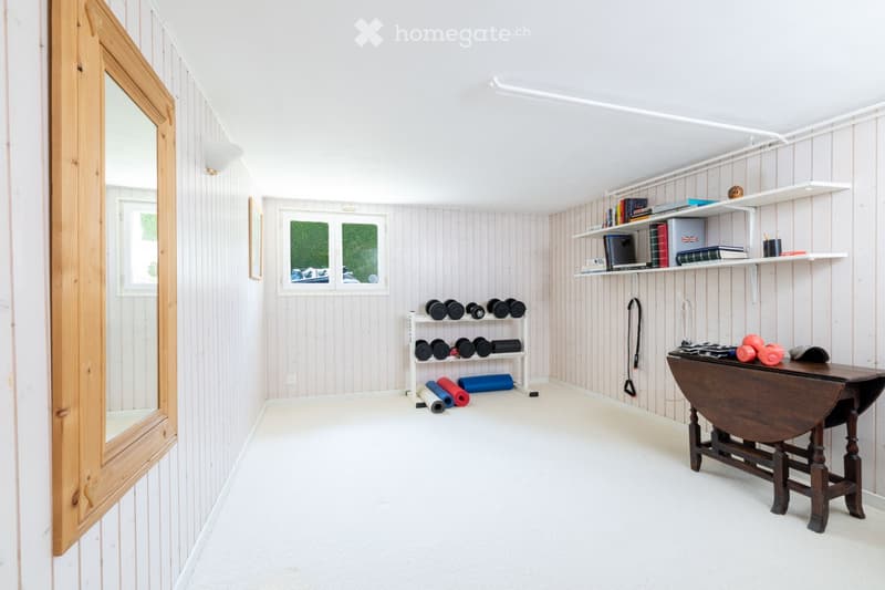 A well-organized home gym with wooden walls, white flooring, and a large mirror. Features include a wooden table, shelves with books and weights, yoga mats, and a white weight rack.