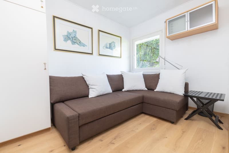 A cozy living room with a brown sectional sofa, white and brown cushions, a small black folding table, a wall-mounted lamp, and built-in cabinets. The room features light wooden flooring and white walls adorned with framed artwork.