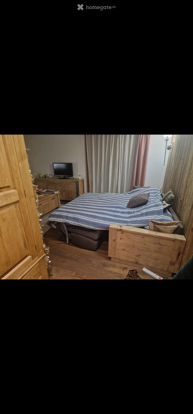 Cozy bedroom with a wooden bed frame, a television on a nightstand, and a curtained window. The bed is neatly made with pillows and a patterned duvet.