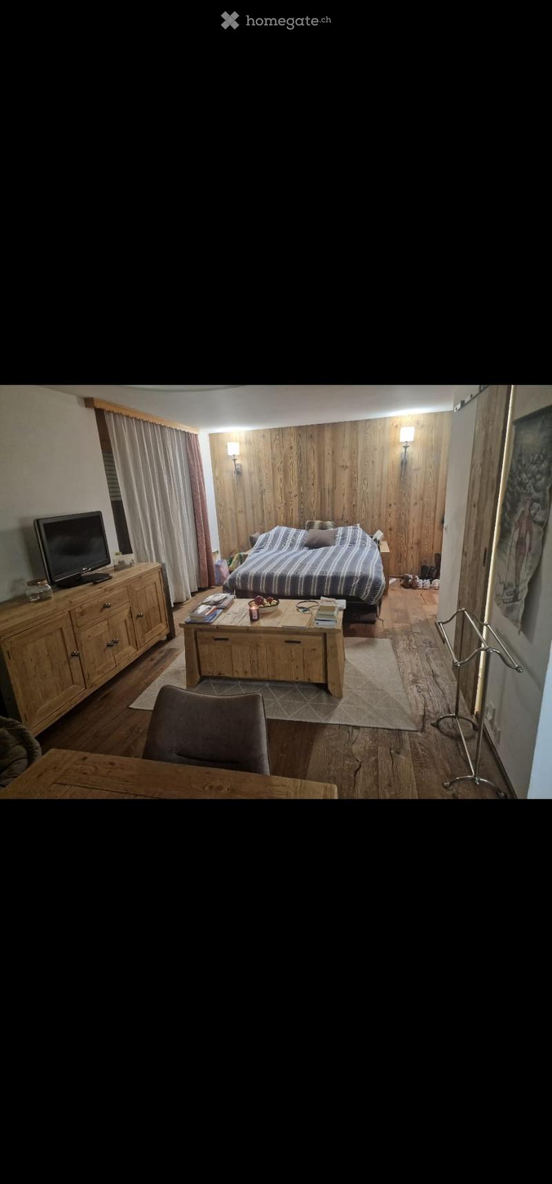 Cozy bedroom with wooden walls, a large bed with striped bedding, a wooden table with a bowl of fruit, a wooden cabinet with a TV, and a chair near the window.