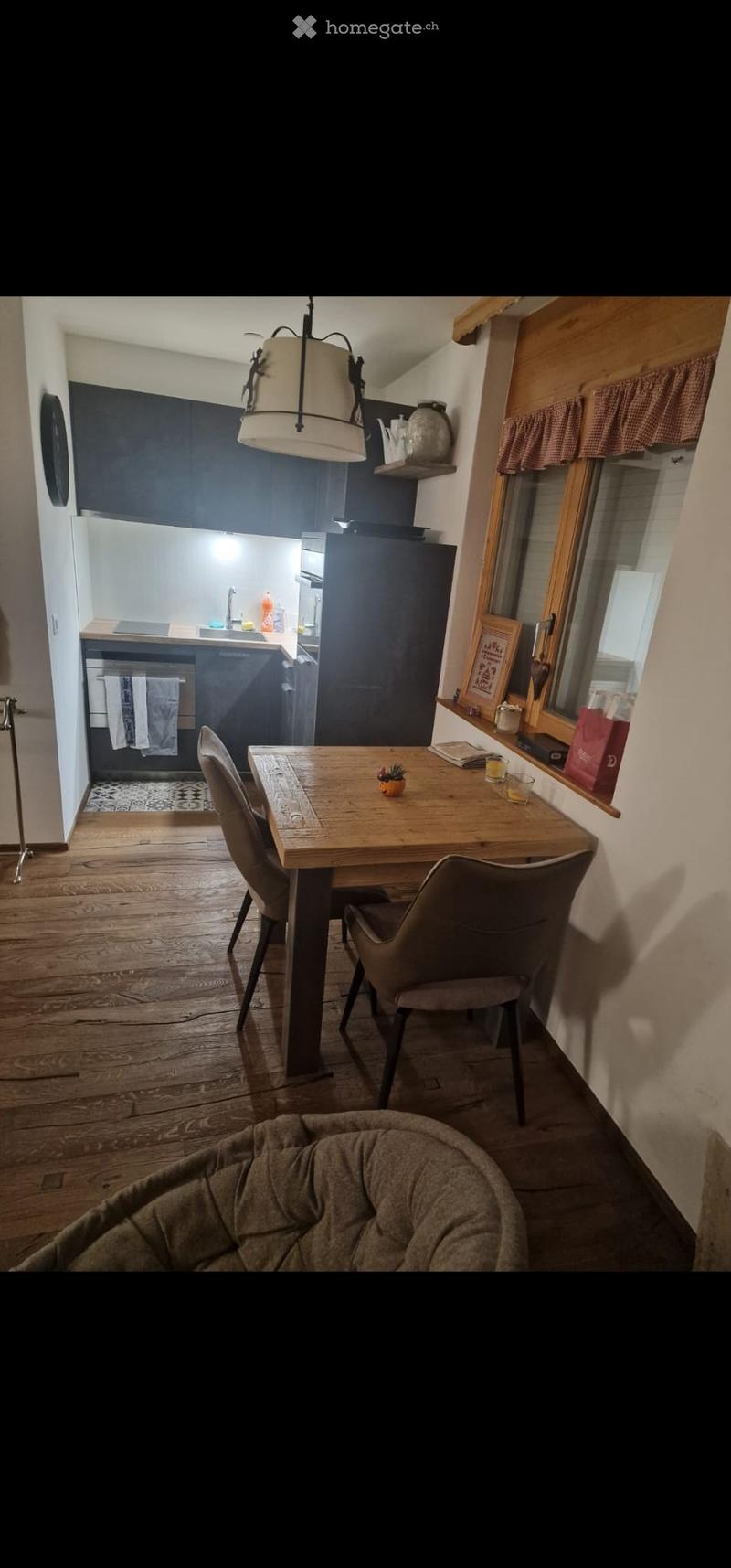 A cozy kitchen and dining area with wooden flooring, a wooden dining table with chairs, a refrigerator, a sink, and various decorative items on shelves and windowsills.
