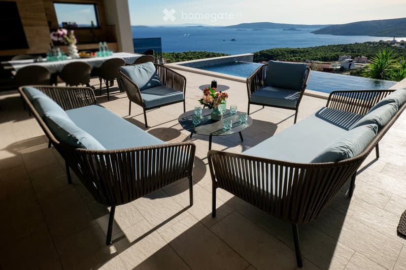 A modern outdoor terrace with wicker furniture, a sofa, chairs, a coffee table, and a view of the sea and distant hills.