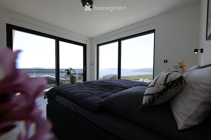 Modern bedroom with large windows offering a scenic view, featuring a bed with pillows, a small table with a plant, and chairs on a balcony.