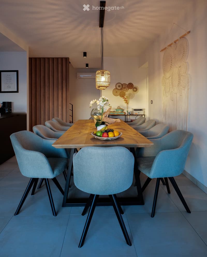Modern dining area with a wooden table, six grey chairs, a fruit bowl, a flower vase, and decorative wall hangings.