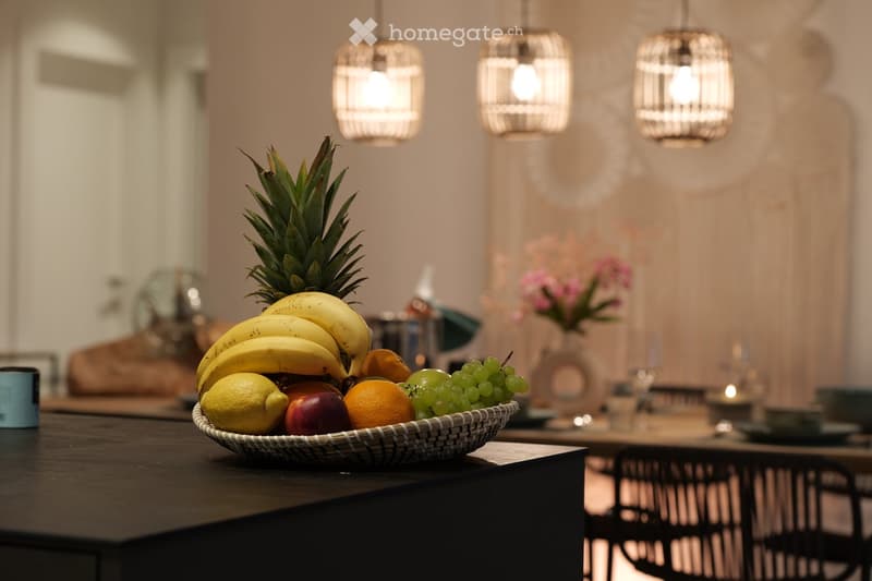 A modern dining area with a dark wooden table, a fruit bowl with bananas, grapes, oranges, and a pineapple, and a vase with pink flowers. There are also wine glasses and a cup on the table.