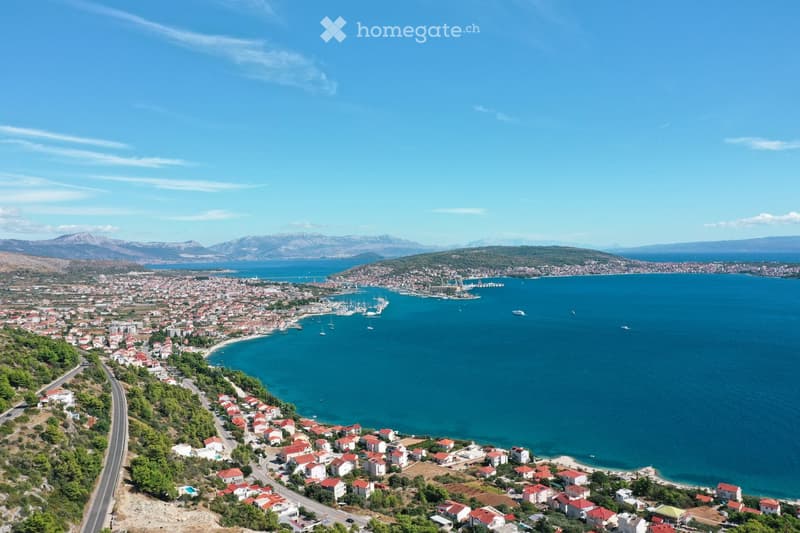 Aerial view of a coastal town with red-roofed houses, a harbor with boats, and a clear blue sea.