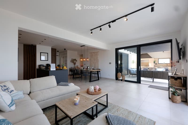 Modern living room with white walls, large sliding glass doors leading to a patio, a sectional sofa, coffee table, dining area with a table and chairs, and a kitchen island with pendant lights.