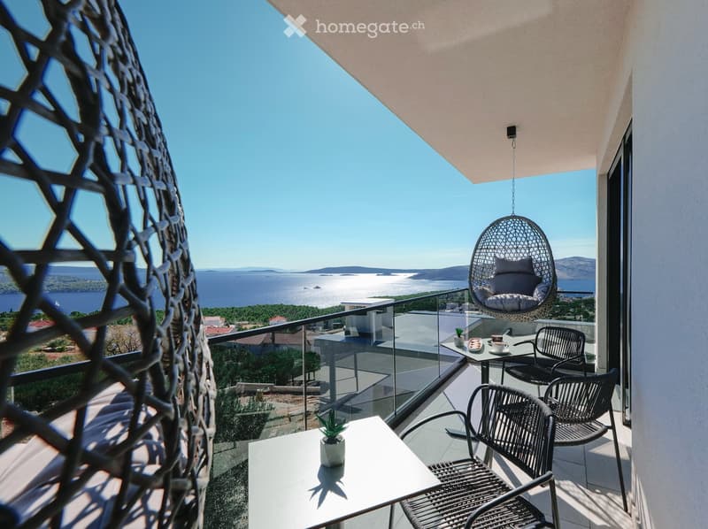 Balcony with a view of the sea, featuring a wicker hanging chair, a small table with a potted plant, and two black wicker chairs.