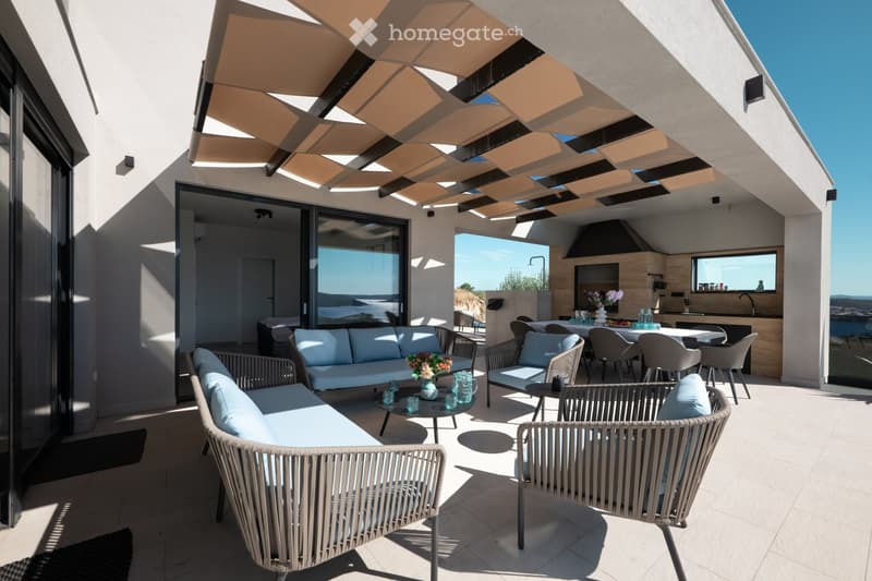 A modern outdoor terrace with a covered seating area, featuring a sofa, chairs, a coffee table, and a dining area with a table and chairs. The space includes a kitchen area with a sink and a view of the sea.