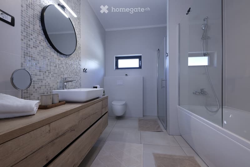 Modern bathroom with a wooden vanity, white sink, toilet, bathtub, and a large round mirror. The space features tiled walls and a glass-enclosed shower area.