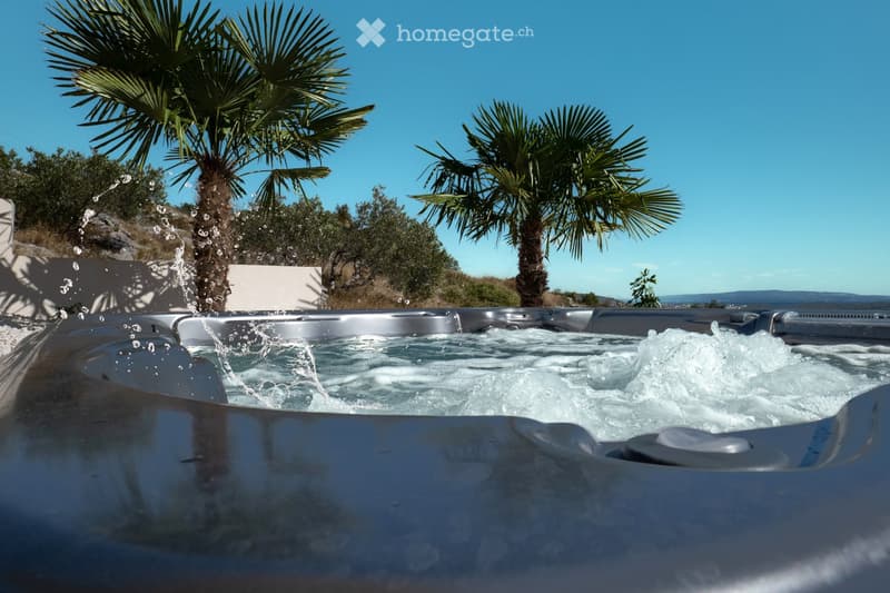 A modern hot tub with bubbling water, surrounded by palm trees and a clear blue sky, with a scenic view of the sea in the background.