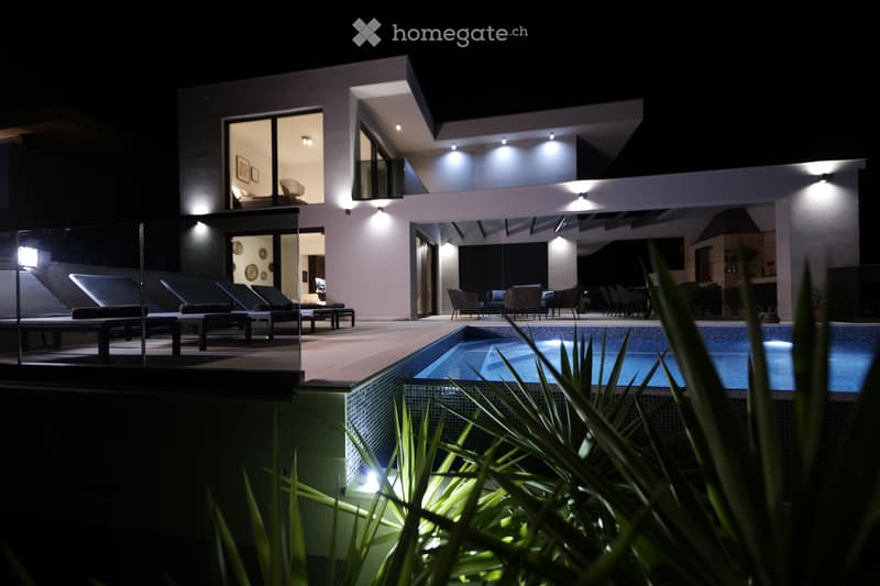 Modern two-story house with a swimming pool, lounge chairs, and outdoor seating area illuminated at night.
