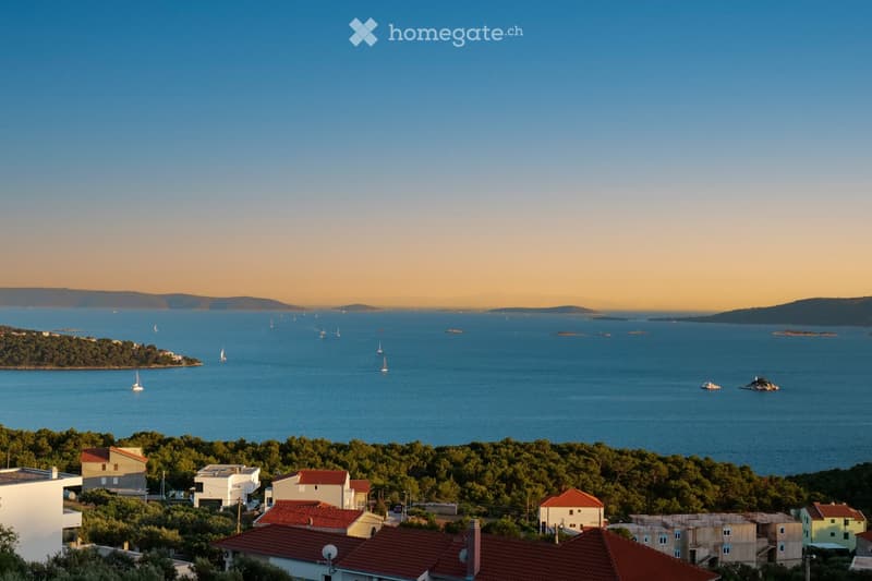 A panoramic view of a coastal town with houses, trees, and a calm sea dotted with sailboats. The sky is clear with a warm sunset glow.