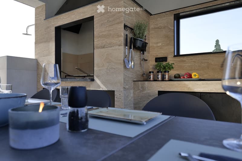 Modern outdoor dining area with a dark wooden table, chairs, wine glasses, candle, and potted plants. Kitchen area visible in the background with utensils and a stove.