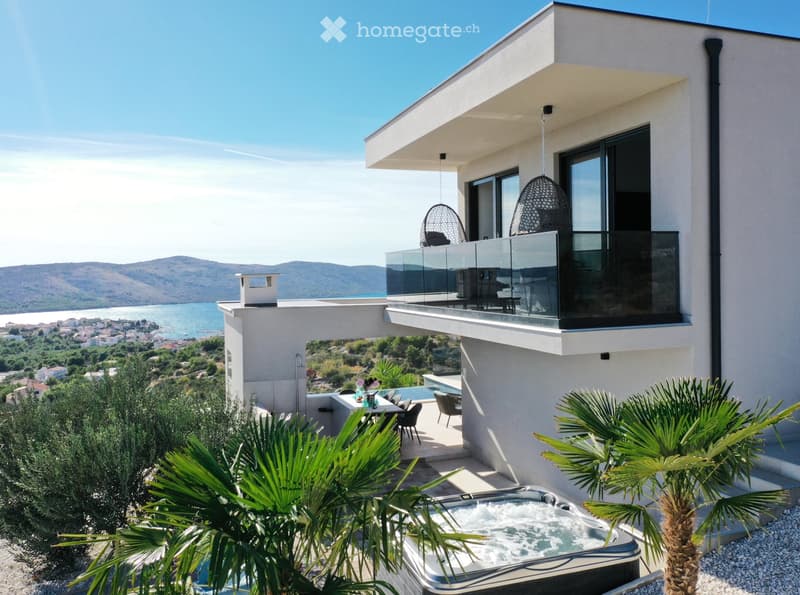 Modern house with a spacious outdoor area featuring a hot tub, dining table, chairs, and a view of the sea and mountains.