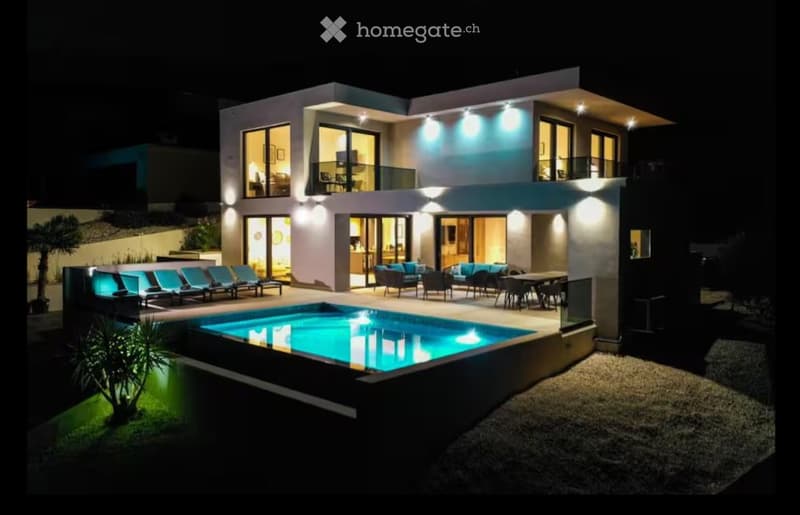 Modern two-story house with large windows, illuminated exterior, swimming pool, outdoor seating area, and lounge chairs.