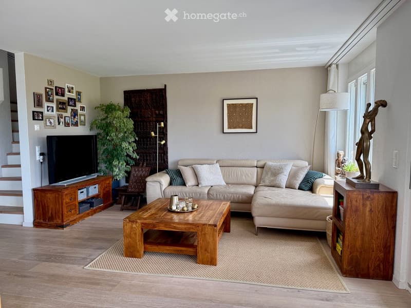 Modern living room with a beige sectional sofa, wooden coffee table, flat-screen TV on a wooden cabinet, floor lamp, decorative picture frame, plant, and a small statue on a side table.