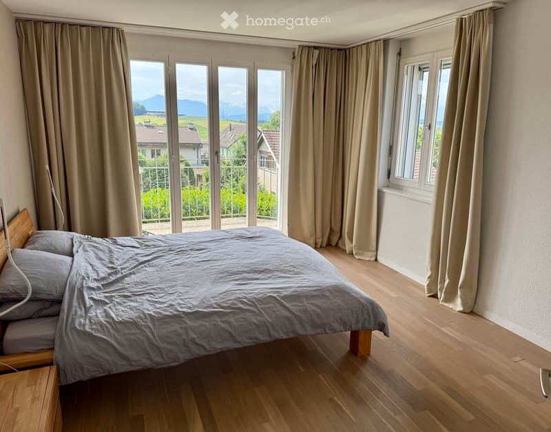 Bright bedroom with wooden flooring, large window and sliding glass doors leading to a balcony, offering scenic mountain views. Features a neatly made bed with grey bedding and a wooden nightstand.