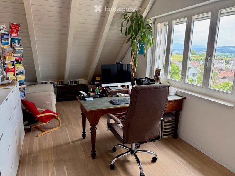Cozy attic office with wooden floors, large windows offering a scenic view, a desk with a computer, a comfortable chair, a small sofa, and a potted plant.