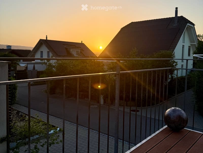Balcony with metal railing, wooden table, and decorative sphere, overlooking a residential street at sunset.