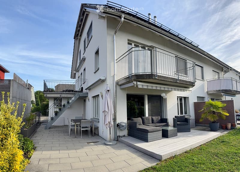 Modern outdoor patio area with a sofa, ottoman, chairs, a table, and an umbrella. The patio is adjacent to a two-story house with balconies and a staircase leading to an upper terrace.