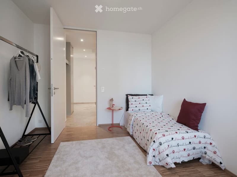 Cozy bedroom with a single bed, decorative pillows, a small round table with a chair, a clothing rack with hanging clothes, and a white rug on the wooden floor.