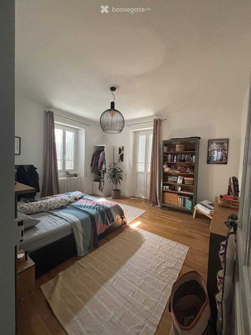 Bright bedroom with wooden flooring, large window with curtains, bed with colorful bedding, nightstand, bookshelf, potted plant, and a small table with a chair.