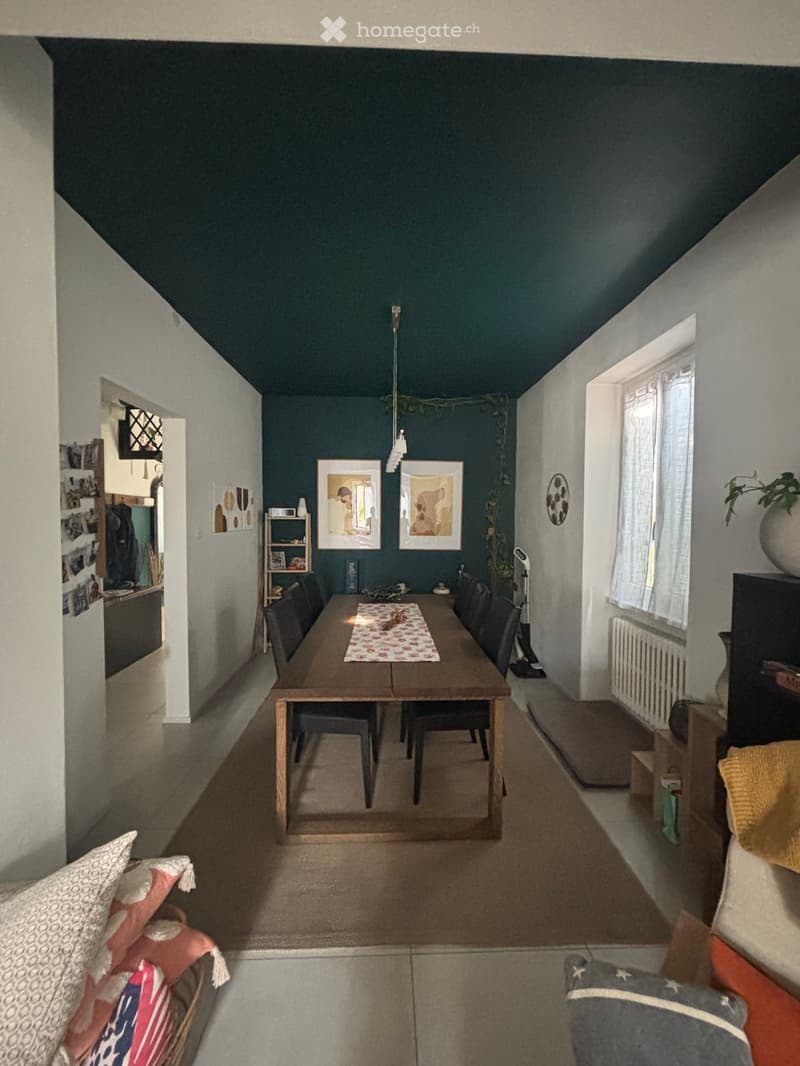 Dining area with a wooden table, chairs, and a sofa in the foreground. The room features dark blue walls, a hanging light fixture, and artwork on the walls.