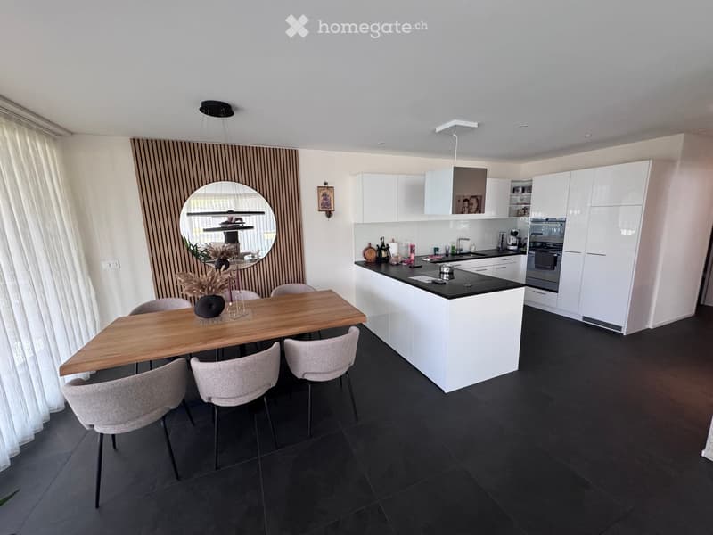 Modern kitchen with white cabinets, black countertops, a wooden dining table, and four chairs. Features a large island with sink and appliances, a round mirror, and a hanging light fixture.