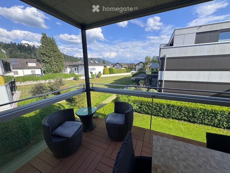 Balcony with wooden decking, glass railing, and outdoor furniture including a table and chairs, overlooking a green lawn and residential area.